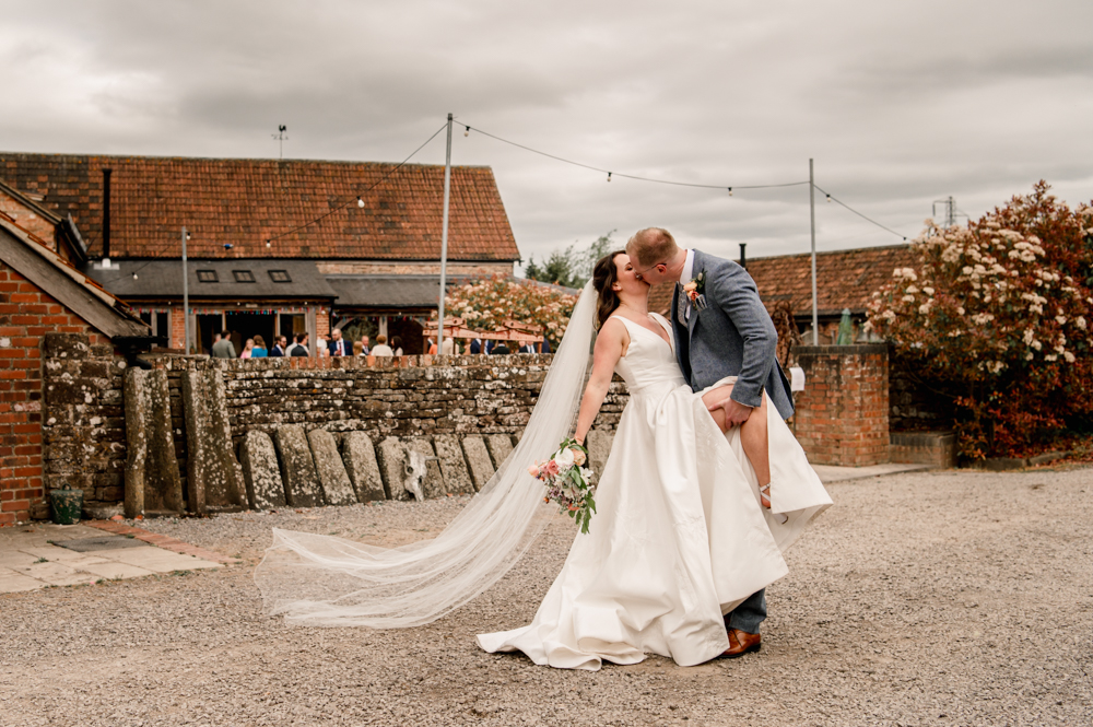 light a bd bright photo of groom kissing bride in a dramatic pose in front of a wedding barn