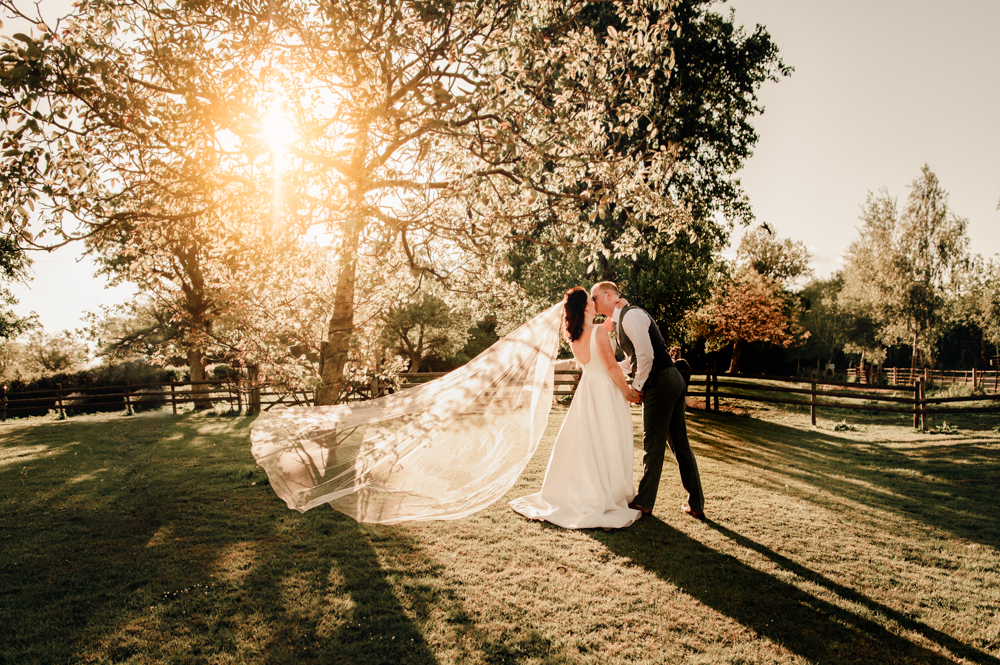 a dreamy romantic shot of the bride and groom kissing under a sun lit tree with her veil flying  