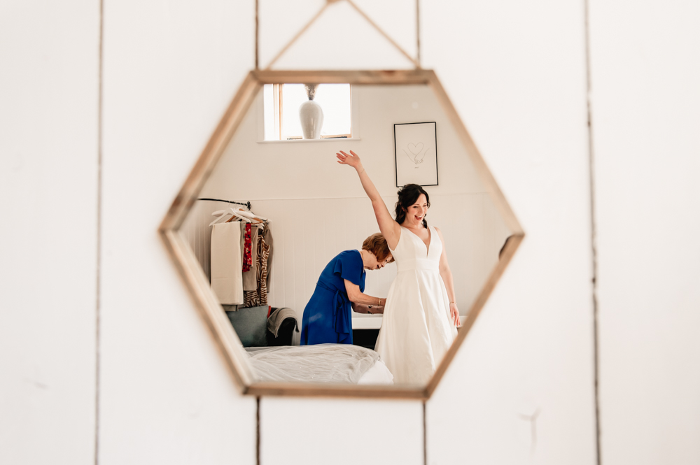 mother of the bride buttoning bride's dress reflection in the mirror