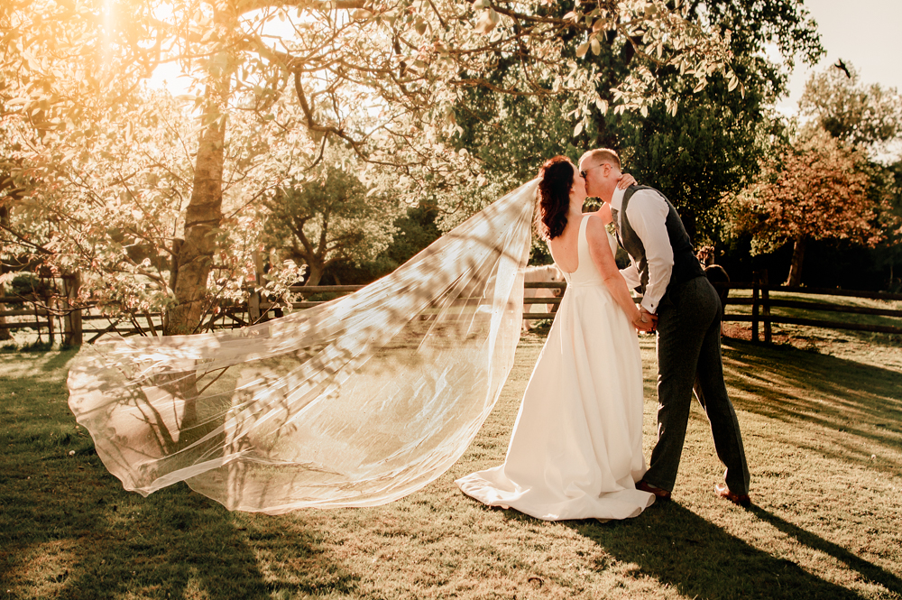a dreamy atmospheric photo of a bride and groom kissing a sun lit forest landscape in the cotswolds with her veil flying and  translucent