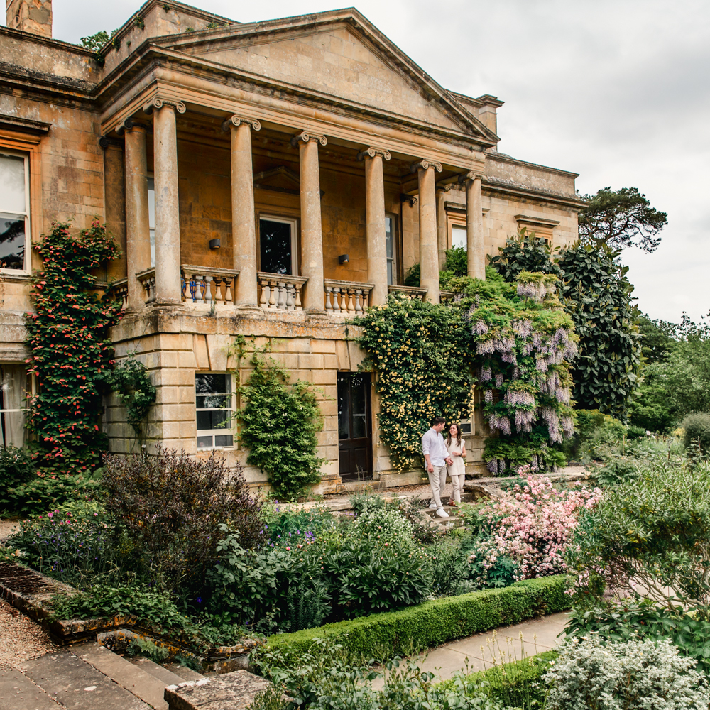 an engaged couple walking in front of a manor house cotswolds 