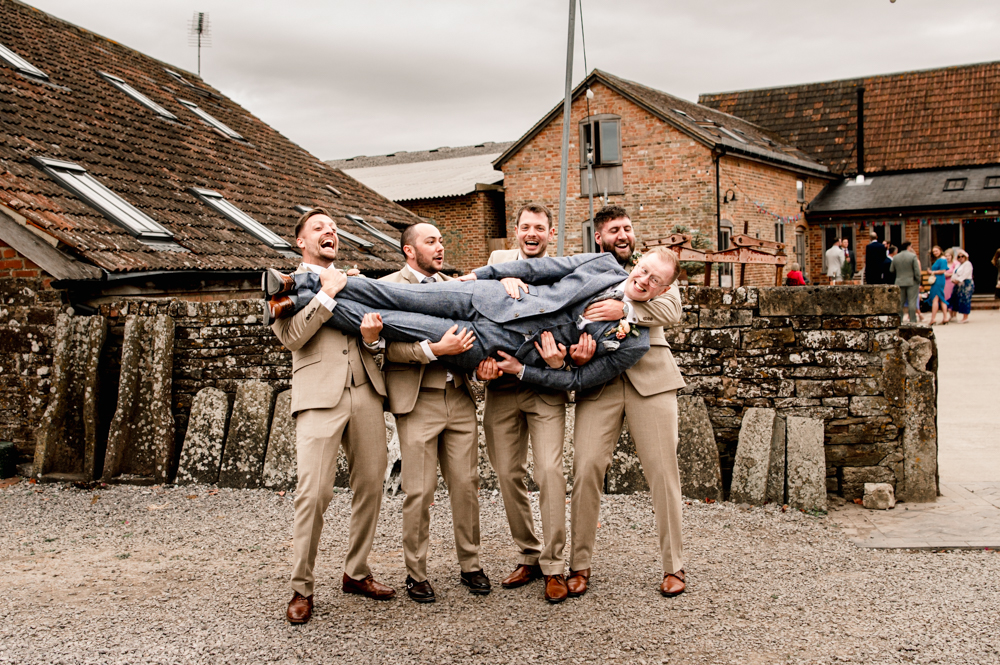 groom being held by groomsmen fun pose with a backdrop is a rustic farm and cotswold stone wall